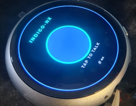 The nyx-badge, first boot — a Waveshare round AMOLED puck showing the indigo-nx badge design: cyan "INDIGO-NX" arced around the top of the ring, a bright cyan core in the centre, "TAP TO TALK" below, "// nx" chip at the very bottom, all against a near-black backdrop, in a black plastic housing on a dark surface